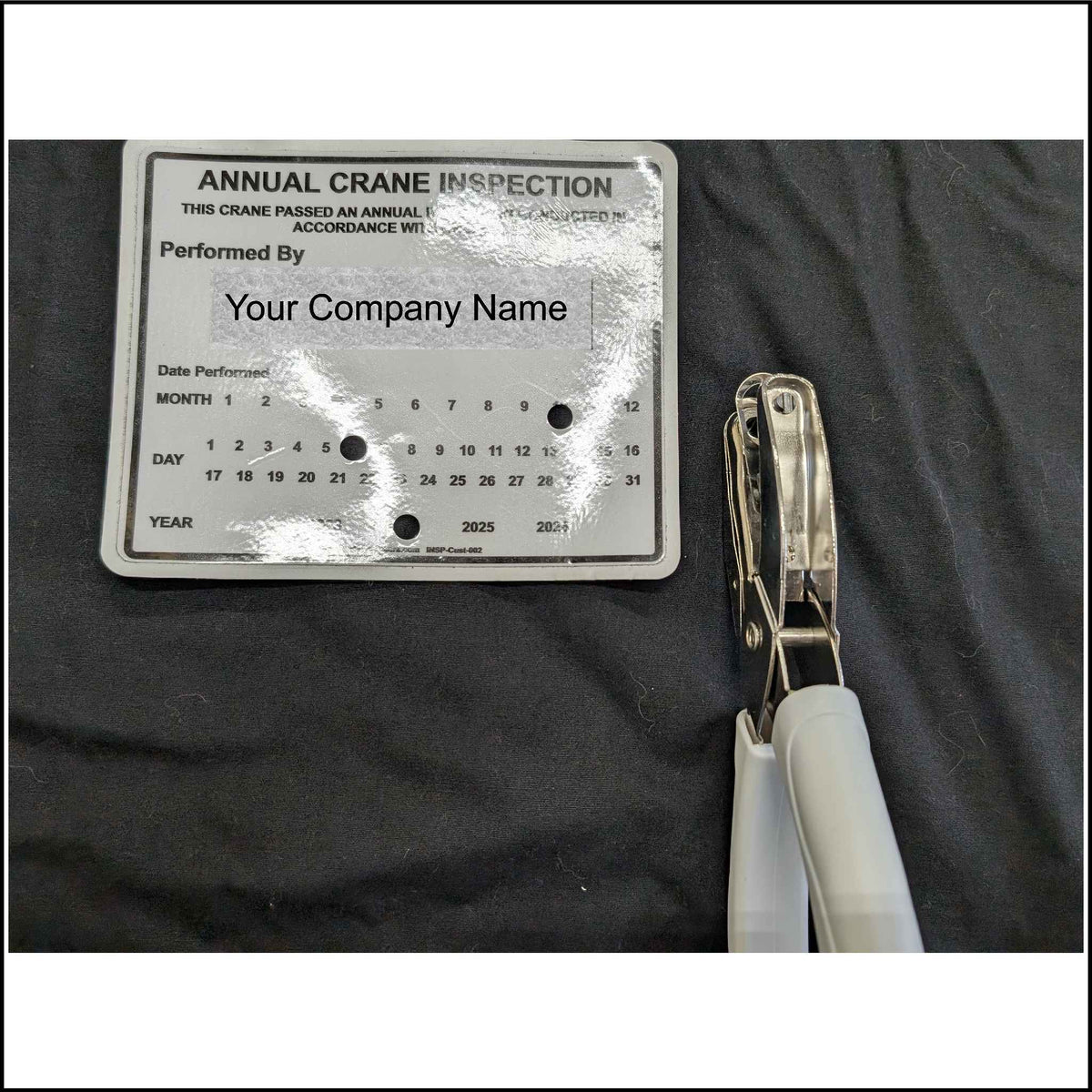 a hole punch with a long reach marking the date on our crane inspection sticker. It is marking the sticker with a 1/4 inch hole on the month, day, and year of the inspection.