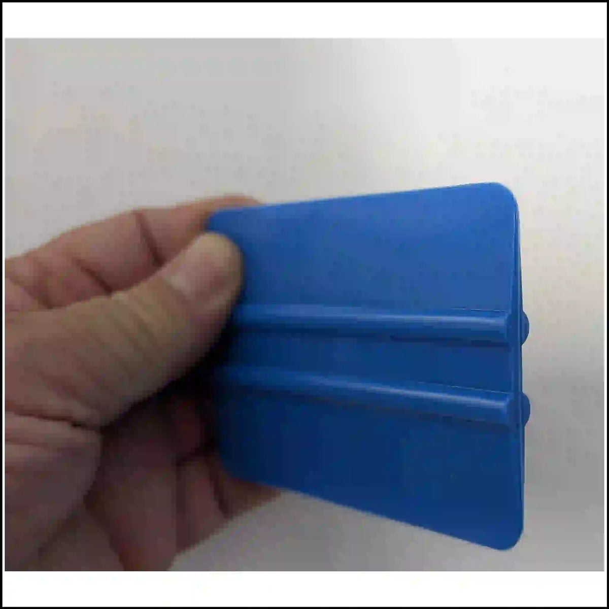 Hand holding a blue decal squeegee at an angle, showing its application edge and raised grip ribs.